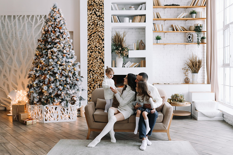 一个幸福的家庭，两个孩子穿着针织毛衣，抱在一起坐在床上庆祝圣诞节，在装饰好的斯堪的纳维亚式室内，冬天的房子的客厅里有圣诞树。图片下载