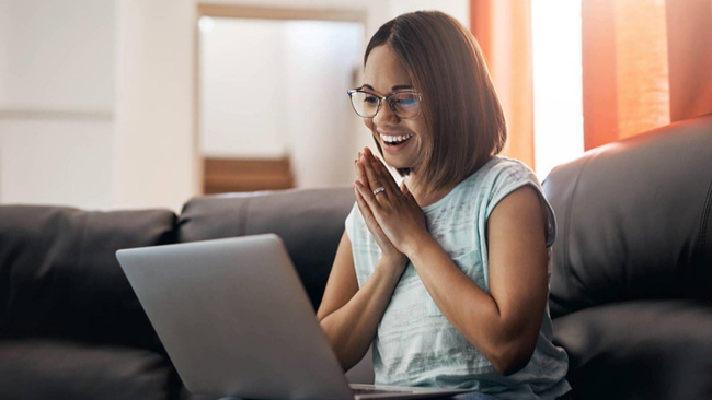 Velocímetro | Mulher sentada em um sofá com o notebook no colo. Ela encara a tela com um sorriso e as mãos juntas próximas ao peito.