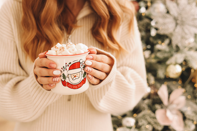冬天的圣诞节，在家里，一位穿着针织毛衣的年轻女子在装饰好的圣诞树前端着一杯带着棉花糖的热咖啡图片下载