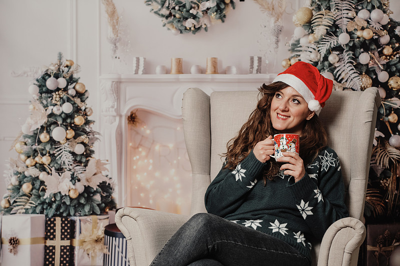 穿着圣诞毛衣，戴着马克杯的女人坐在扶手椅上抬头看图片下载
