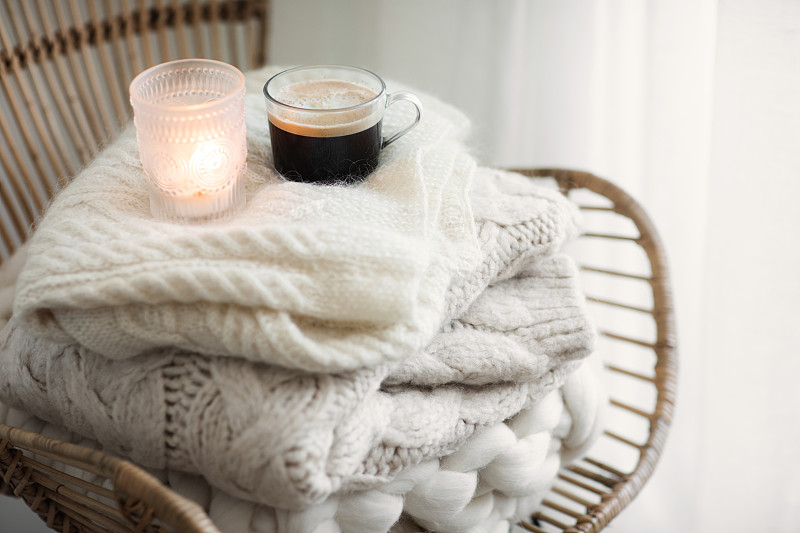 一堆舒适的白色羊毛针织毛衣，柳条扶手椅上放着蜡烛和咖啡。Hygge风格。温暖的概念。图片下载